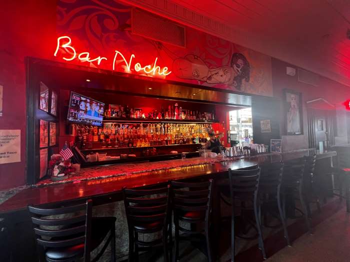 A bar with a long counter, lots of bottles on the shelves behind the counter, and a red neon light saying Bar Noche!