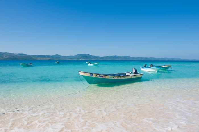 small blue and white fishing boats on the crystal clear waters outside a crispy white sandy beach under blue skies