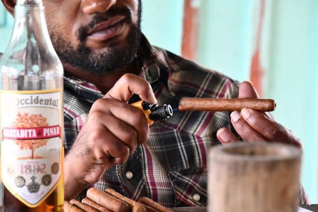 A man in a flannel shirt lighting a Cuban cigar next to a bottle of rum