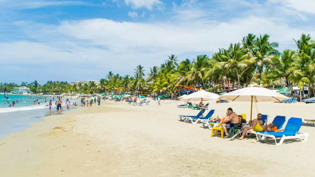 A warm sunny day at the white sandy Cabarete Beach in the Dominican Republic, with lots of people enjoying themselves on the beach and on sun beds under parasols along the green palmy beach shore