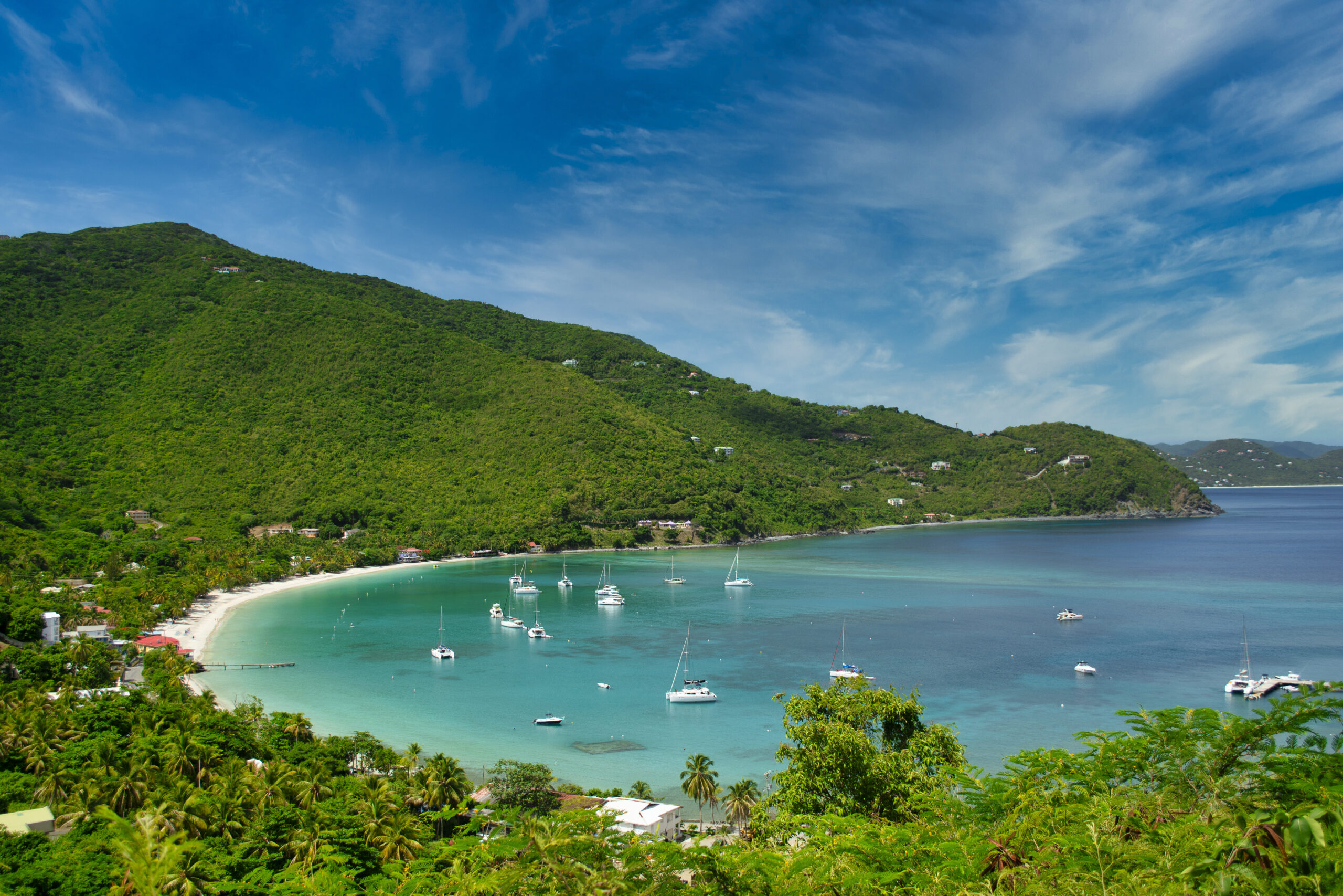 Cane Garden Bay on Tortola, a secluded bay with a thin sting of sandy beach in front of the lush green forest and hills, and boats on the greenish blue water