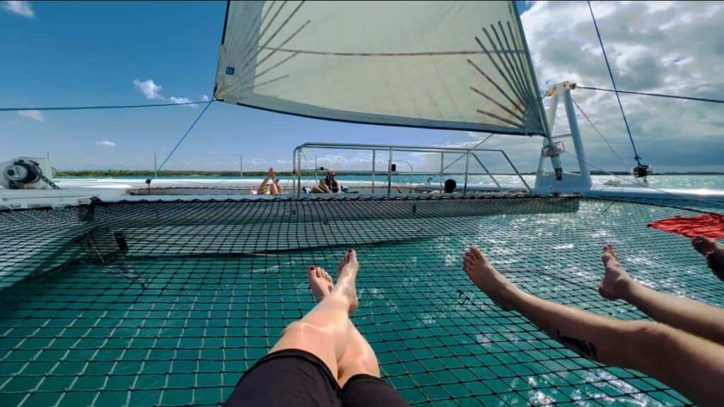 Two people chilling on the net of a catararan, only legs visible, with the white sail above, and greenish water below on a sunny summer day