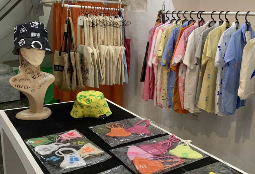 A table in a shop covered with items for sale like caps, bags, and shirts on a rack in the background