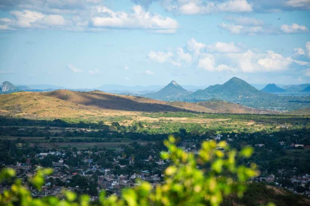 An open landscape with small houses nestled among trees, wide green plains, and green hilltops in the distance under a blue sky in Cuba