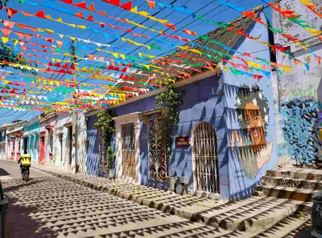 A charming street with small old houses in different colors with stunning architectural details on doors and windows, murals, and colorful decorations overhead under a deep blue sky. 