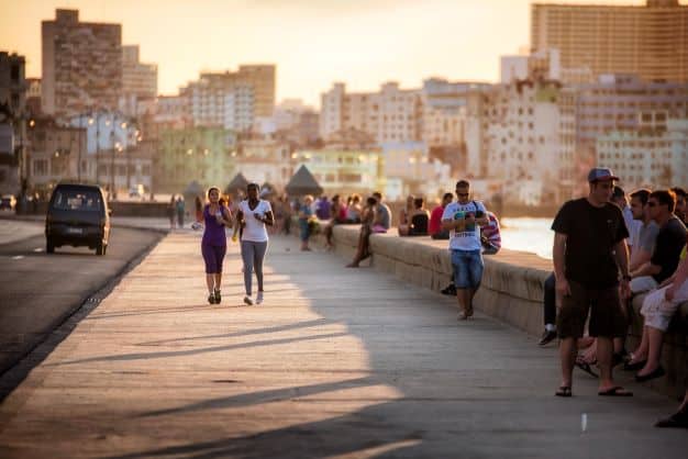 A boardwalk along the sea on a sunny day with a rugged city skyline in the background and lots of people walking and sitting on the boardwalk