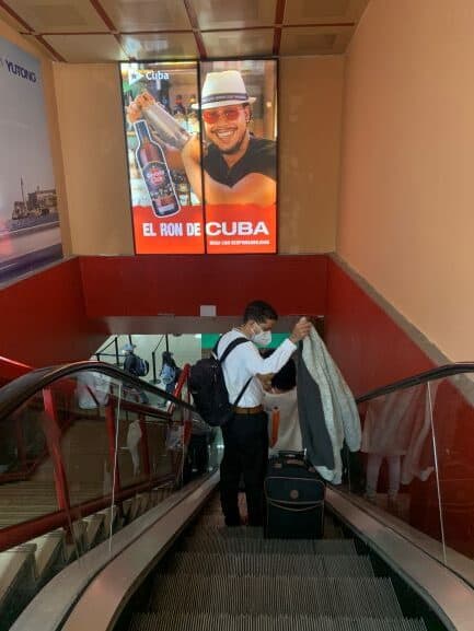 A man in an escalator in the airport in Havana, Cuba, wearing a face mask, under a big commercial for Cuban rum