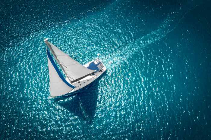 A white sail boat with bright white sails on a shiny blue sea seen from above