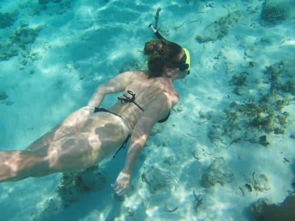 A woman snorkeling underwater, with a black bikini, in crystal clear waters with the white sandy bottom underneath