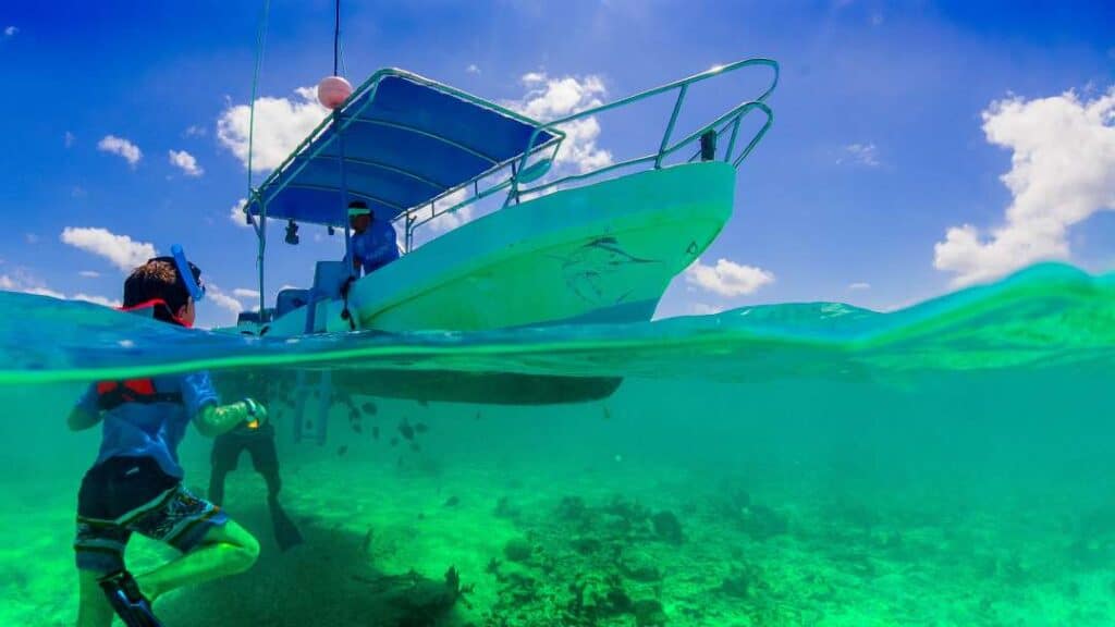 Snorkeling from a boat, photo halfway over and halfway under water, a boy in the water net to the blue and white boat under a blue sky, with the white sandy bottom and marine life under the surface