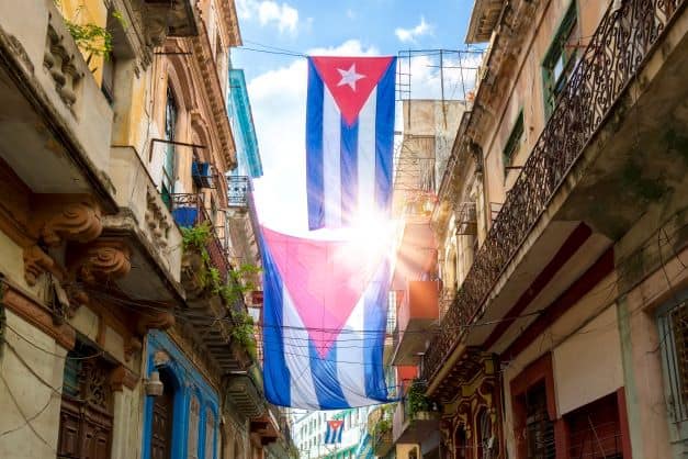 A narrow street in Old Havana with a Cuban flag hoisted between the buildings. The sun shining from behind through the flag. Interesting facts about Cuba
