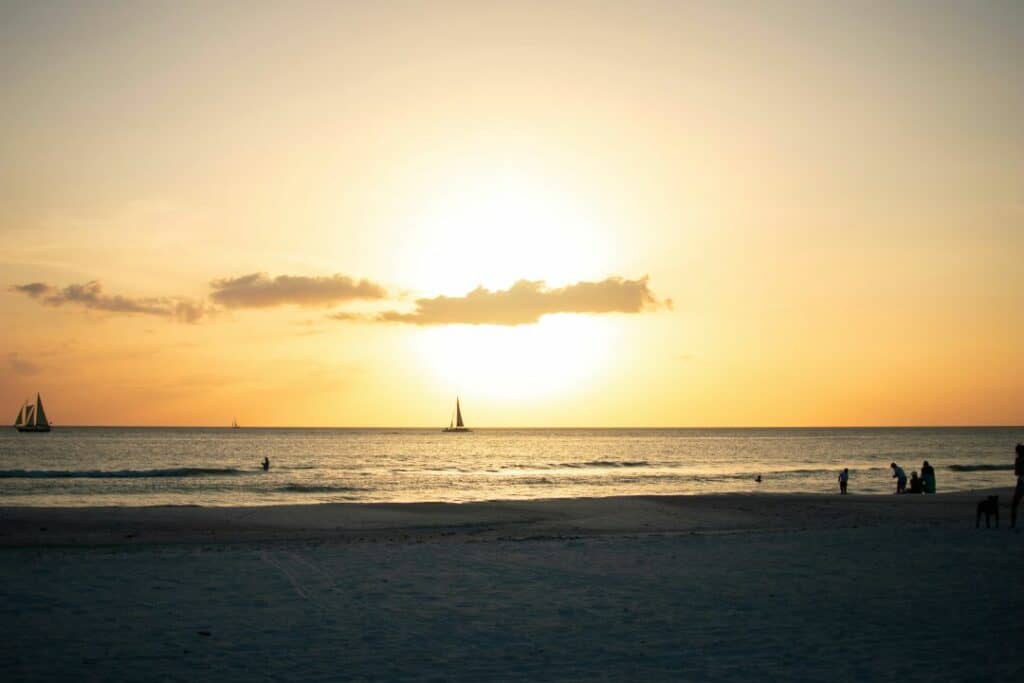 Glowing sunset over a white sandy beach with people still in the surf, and a sailboat in the horizon