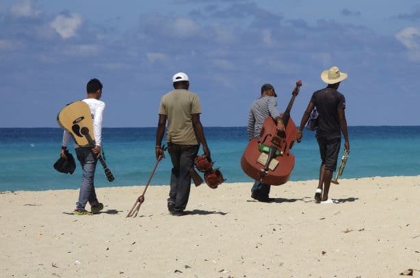 How to get a tourist card for Cuba. Musicians walking along a white sandy beach with their instruments, on a bright sunny day.