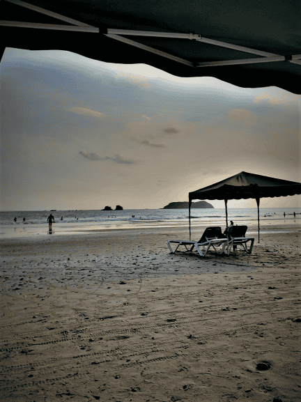 A sandy beach at low tide, with people far away in the water under a pale blue sky