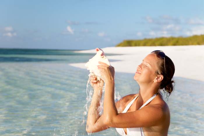 Why worry about hurricanes in the Caribbean? Woman sitting in the clear warm water on the beach, smiling and looking up at the sun, and holding a sea shell. 
