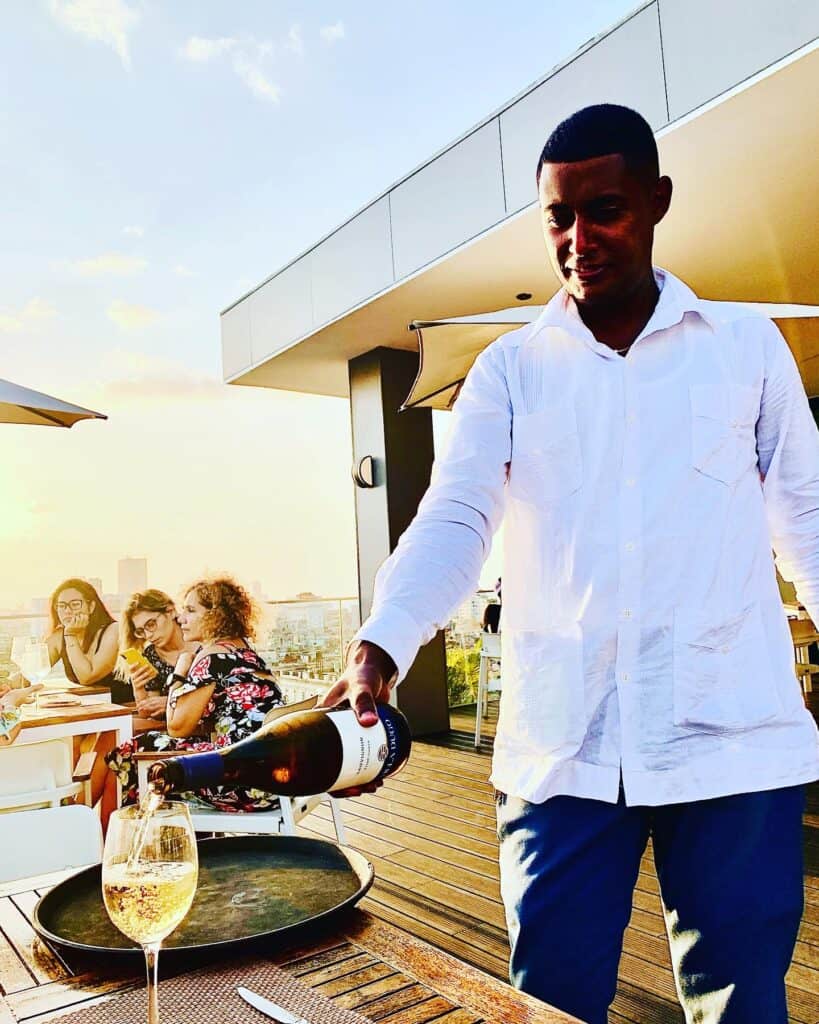 A waiter dressed in white pouring white wine in my glass on a rooftop restaurant in Havana Cuba. It is a bright summer day, and the sun is shining. 