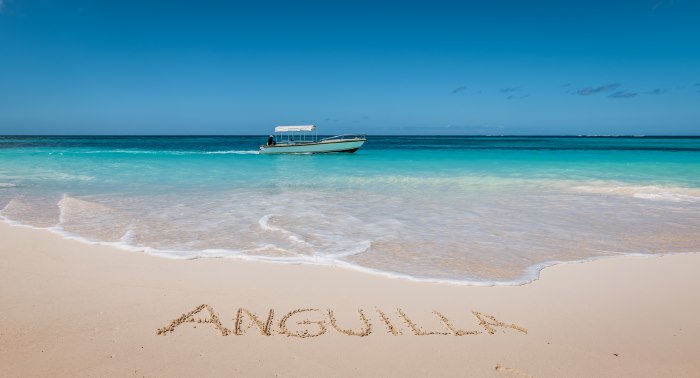 Paradise golden sandy beaches in Anguilla, with the word "Anguilla" written in the sand in front of the crystal clear bluish water with a white boat passing a few yards from the shore