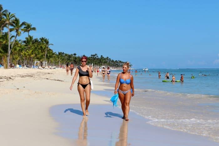 Two women walking along Uvero Alto beach in PUnta Cana, the Dominican Republic. The sand is light yellow, palms border the sands, and there are lots of people in the water on a warm sunny summer day. 