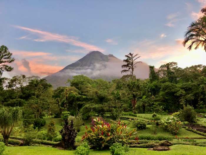 The impressive Arenal Volcano outside San Jose on a bright summer day right before sunset, the volcano top is surrounded in floating transparent clouds, the higher clouds have a glow from the and in the front of the photo is a lush green park with plants, beautiful flowers and trees. 