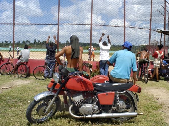 A baseball match in Cuba, played inside a tall fence, and random people watching from the oudside sitting on bikes and even a motor bike, on a bright summer day. 