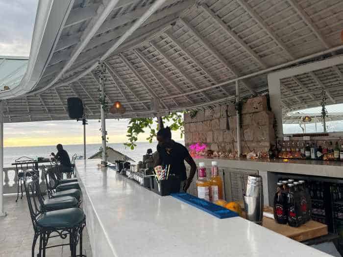 Elegant beach bar in Montego Bay at sunset, with a long, white bar, stone architecture behind the bar, and the ocean in the distance