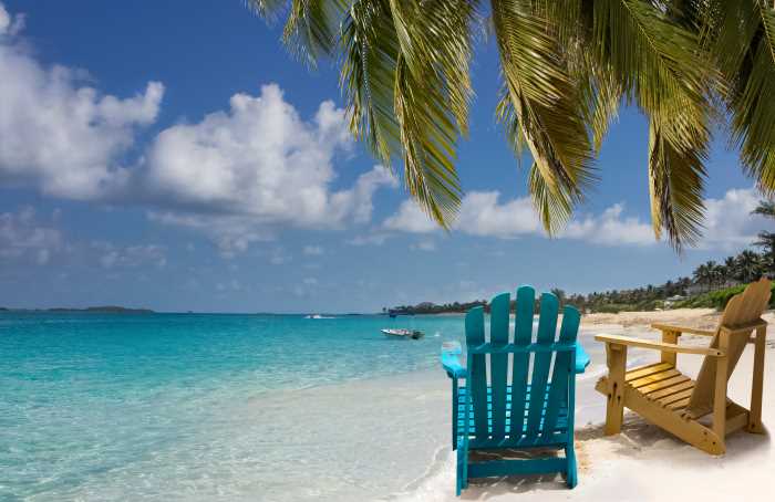 Paradisiacal white beach in Nassau, the Bahamas, with beach lounge chairs sitting IN the water on the white sandy beach under the palm trees next to the crystal clear ocean