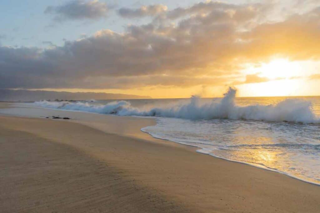 Incredible beach at sunset in Oahu, Hawaii, with golden sands, whitewater coming in with big waves, and a bit of golden clouds in the horizon where the sunset in shining through