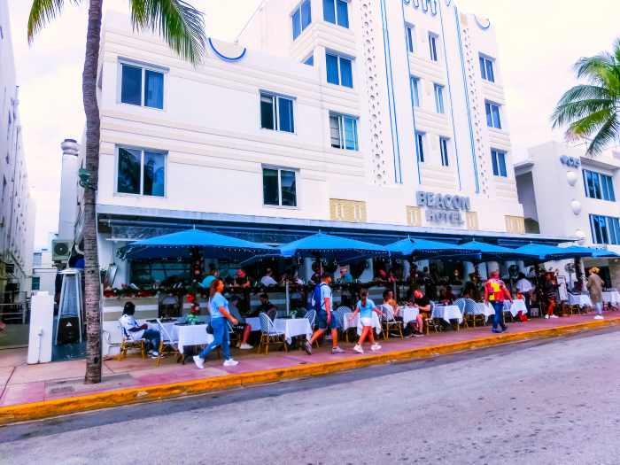 The white facade at teh Beacon Hotel on Miami Beach, with blue details, and outdoor dining areas with white clothed tables along the sidewalk. 