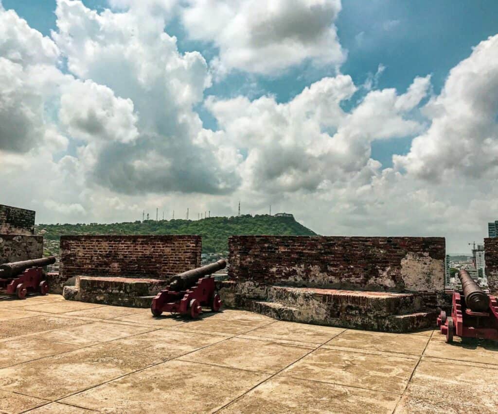San Felipe Fortress with old stone structures, and two old cannons still pointed at possible enemies. The green hills in the background under the blue partly clouded sky. 