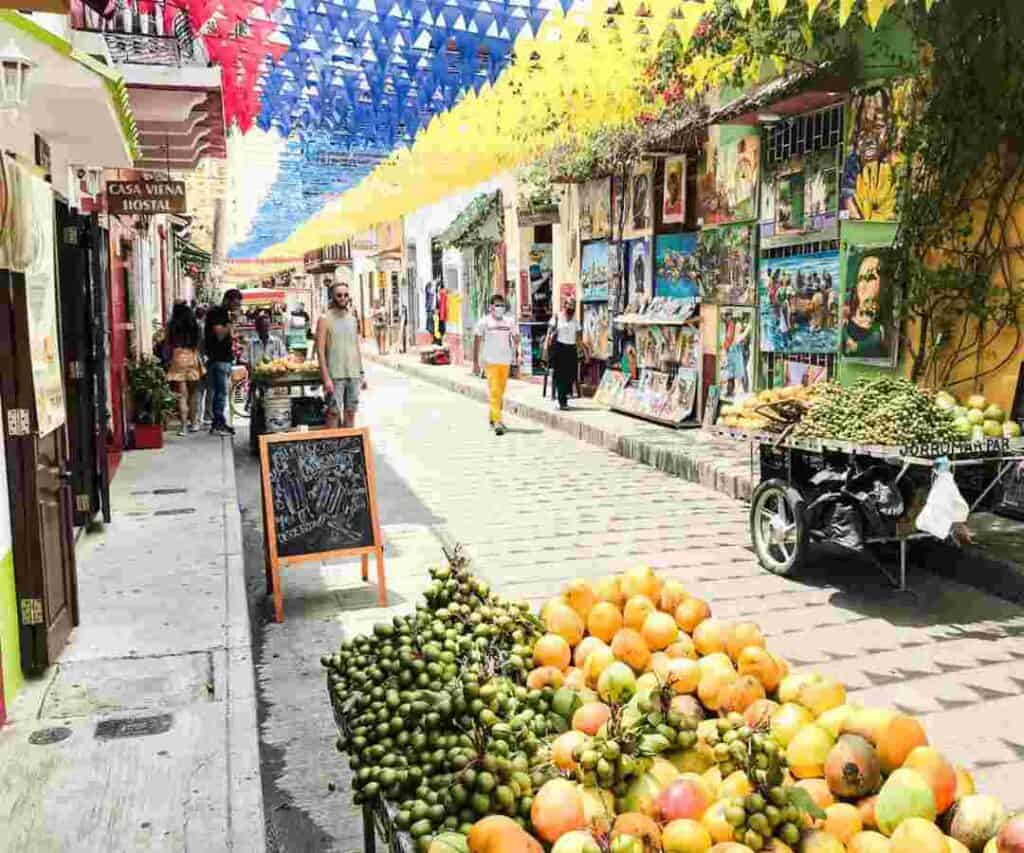Street ambiance in Cartagena, a cobblestoned street with fruit carts, colorful art for sale, cages, shops and people under a partly shading ceiling of yellow and blue decoration all along the street