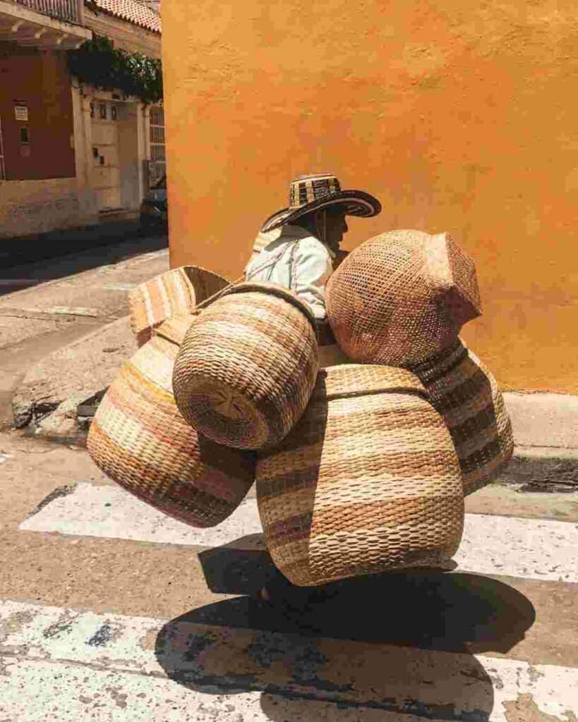 Basket salesman in Cartagena loaded with large beige baskets, wearing a cowboy hat, walking along a street with orange walls in the sunshine