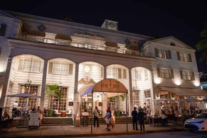 The charming facade of the Beysy Hotel on South Beach at night. White typical southern architecture, a wooden building with charming details, tall arched entrances to the wooden porch, large windows and an inviting ambiance with lighting and life. 