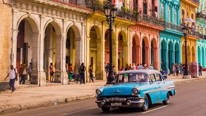 Pastel colored beautiful colonial buildings along a street in Havana, with tall archways, columns, and people on the sidewalk. A blue classic American car in the front of the photo. 