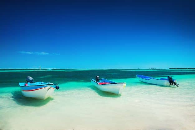Three small blue and white boats lying in the crystal clear waters on the beach in the Cuban Keys, the water is greenish and beautifu., and the sky above is bright blue. 