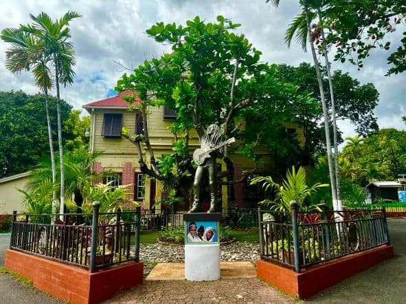 The entrance to Bob Marleys house, which is now a museum. The entrance is a small opening in a black iron fence based on red brick, and inside is a beautiful garden an a large tree in front of the yellow house. 