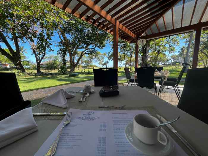 The white breakfast table with a cup of coffee at the Blue Jacktar in Puerto Plata, with open views to the garden with trees and green grass on a bright sunny day. 
