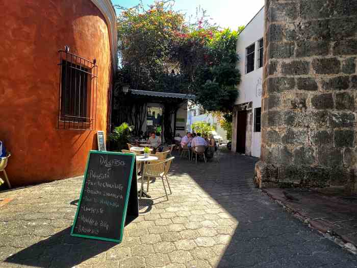 Charming coffee shop in Zona Colonial, with a warm orange colored building on the left, old stone structures on the right, and an intimate outdoor seating area with green plants and bushes