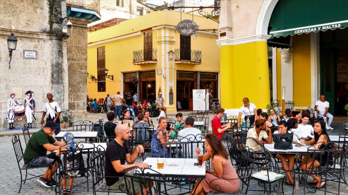 Outdoors cafes on Plaza Vieja in Old Havana Cuba. Colorful classic colonial buildings encircle the cozy plaza with charming seating areas where lots of people are enjoying the day. 