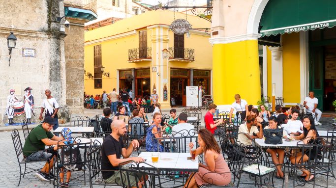 Charming outdoor seating in Plaza Vieja in Old Havana. Small tables with decorated chairs, alongside a yellow brifh building and colonial architecture. 