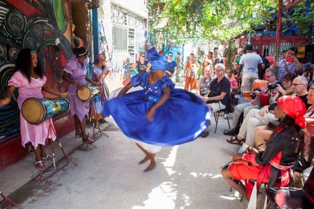 Colorful dancers in Cuba, a woman dancing fiercely wearing a bright blue wide dress and headband while two others play the drums in front of seated impressed spectators. 