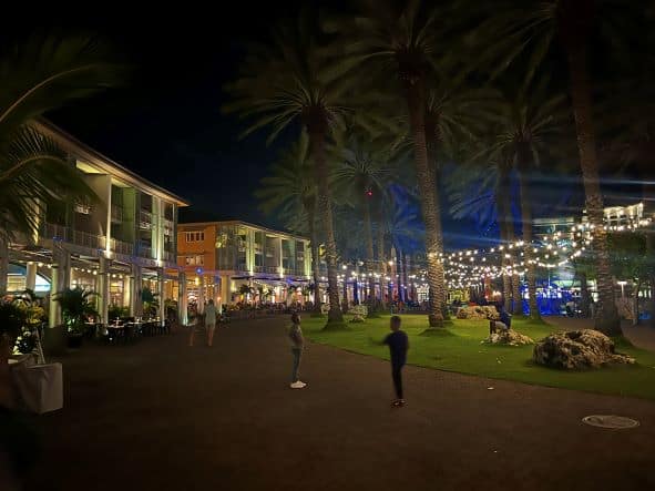 Camana Bay at night, with restaurants like beads on a chain, beautiful lighting against the dark sky, lights hanging between the palm trees in the park area, and lots of people enjoying the evening. 