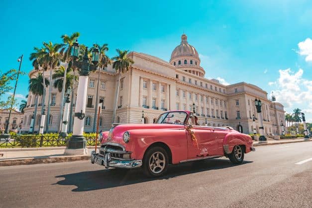 The grand white building of the Capitolio on a bright summer day in Havana, with a bright pink classic American convertible car in front. 