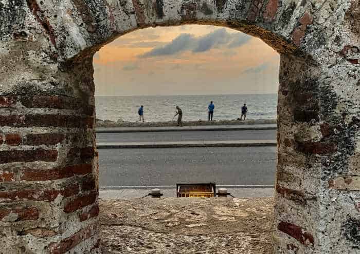 You can peek out through the Old City Wall in Cartagena through old arched holes, and here you can spot four fishing men outside on the shore trying to get the catch of the day in the late afternoon. 