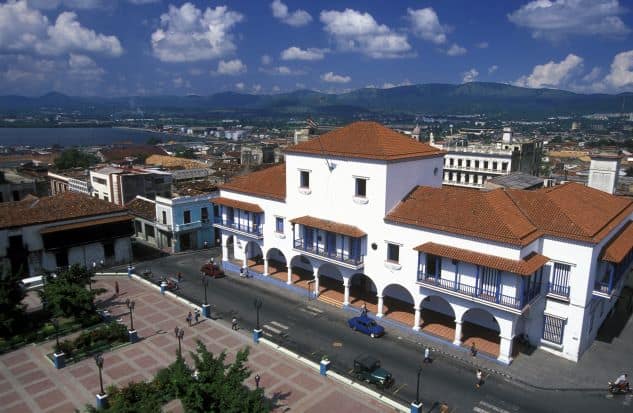 The white colonial casa de Velazques in Santiago de Cuba, next to Parque Cespedes, a large building with red stone roofs, and the city is in the background. 