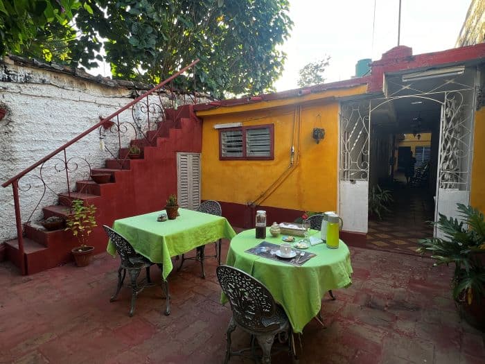 Casa particular breakfast in a charming backyard in Trinidad, tables with green table cloth, orange house with white doors, and a red brick stairwell to the terrace. 