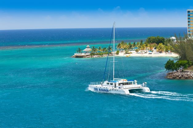 An elegant white catamaran heading out to sea on the clear blue water outside Ocho Rios, passing a white sandy beach strewn with palm trees. 