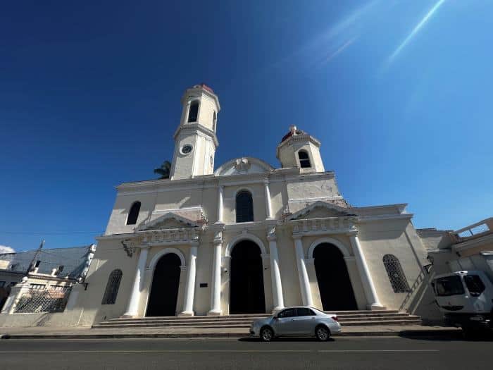 The white cathedral in Cienfuegos with tall arched doors, and two white towers in different heights, facing the main plaza in the city on a bright summer day with blue skies behind it. 