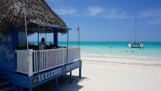 A blue and white beach bar sitting on the wihte sandy beach in Cayo Coco in front of the crystal clear blue water with a white catamaran on a sunny day with blue skies