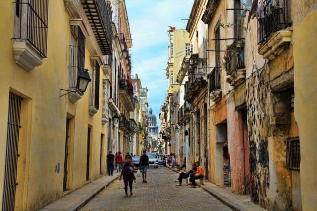 Narrow street in Central Havana, with colonial houses on both sides in different shades of yellow. Many facades are run-down, including the small elegant balconies, and there are people walking and resting in hte street. The majestic Capitolio can be seen far away in the background of the photo. 