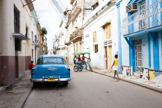 A typical street in Central Havana, each classic colonial house has its own color, the street is rugged, locals walking, a bicycle taxi on its way with passengers, and a blue classic American car is parked on the curve. 